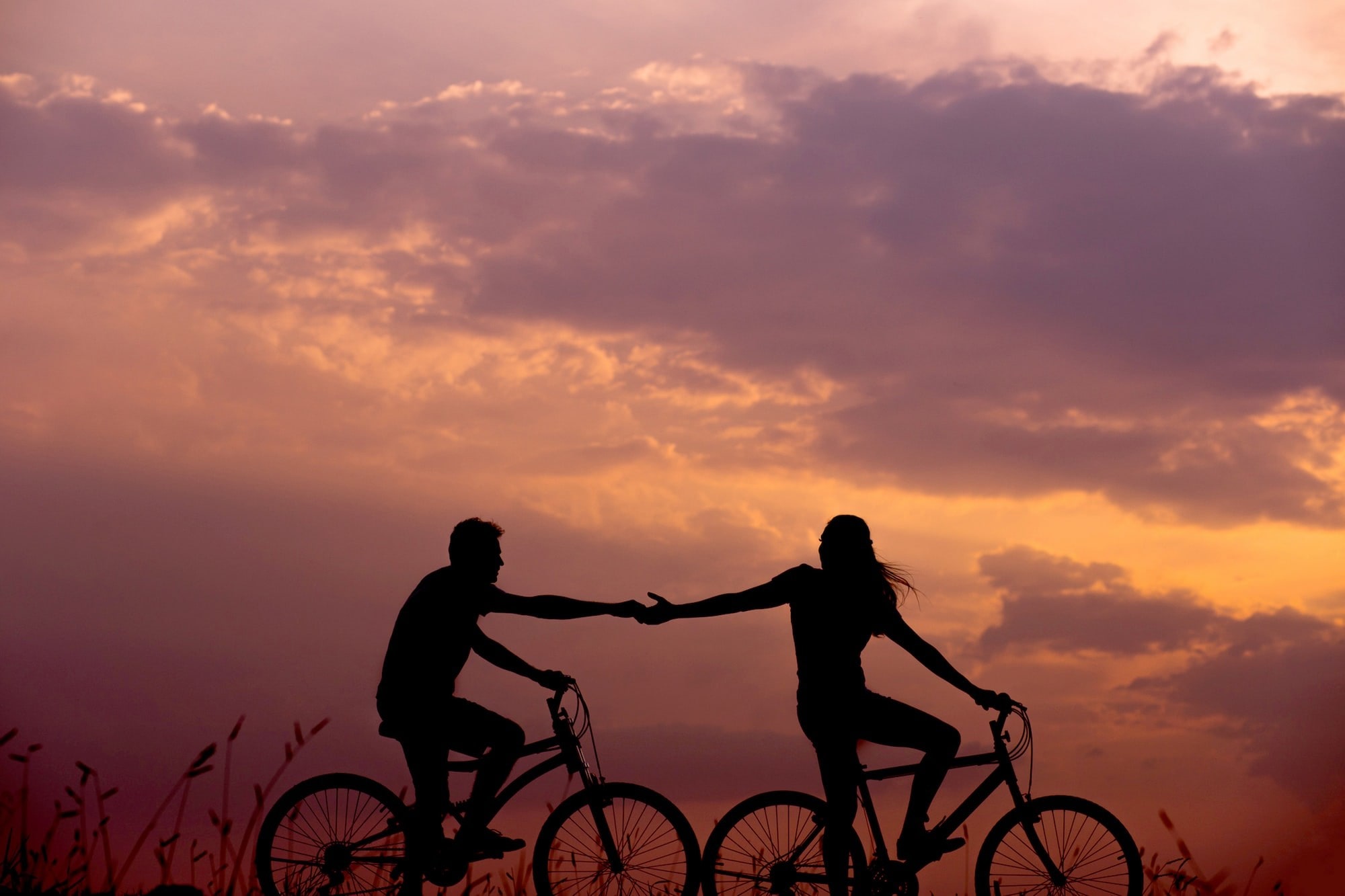 Image of two people riding bicycles. The person in front is reaching back to the person behind her to help and connect with him.